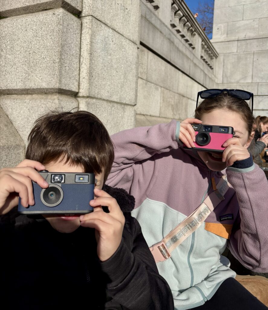 Two children holding Camp Snap cameras and smiling while sitting outdoors.