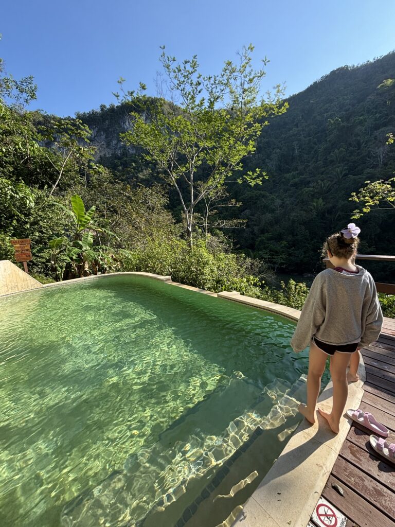 A child stands by the edge of a serene swimming pool surrounded by lush greenery and mountains in the background.