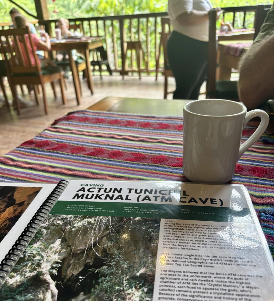 View of a cozy dining area at Black Rock Lodge with a menu displaying an adventure tour for Actun Tunichil Muknal cave, a coffee cup, and vibrant woven table decor.