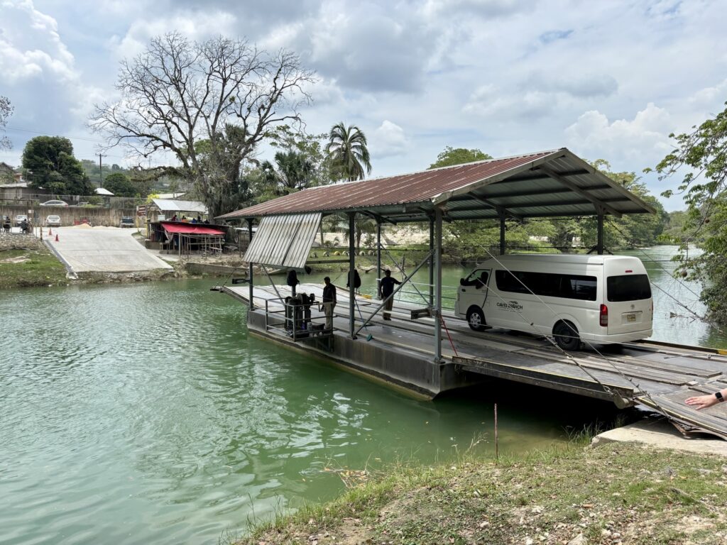 A hand-cranked ferry transporting a van across a river in Belize, surrounded by lush vegetation and a clear blue sky.
