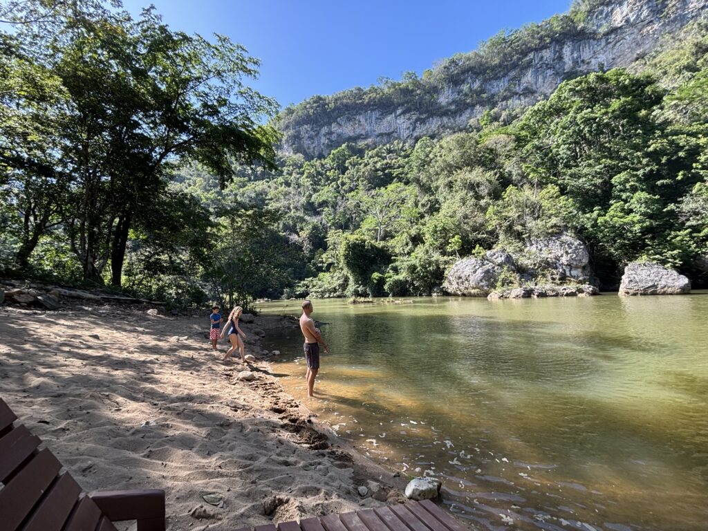 Family members wading in the shallow waters of the Macal River at Black Rock Lodge, surrounded by lush green hills and trees.