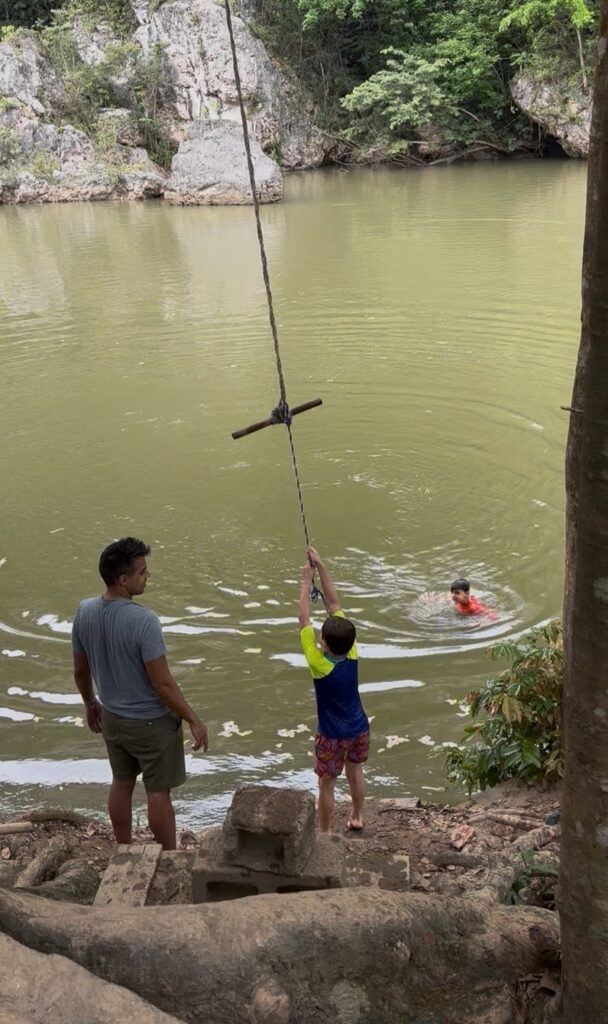 A child prepares to swing from a rope into a green river while an adult looks on nearby.