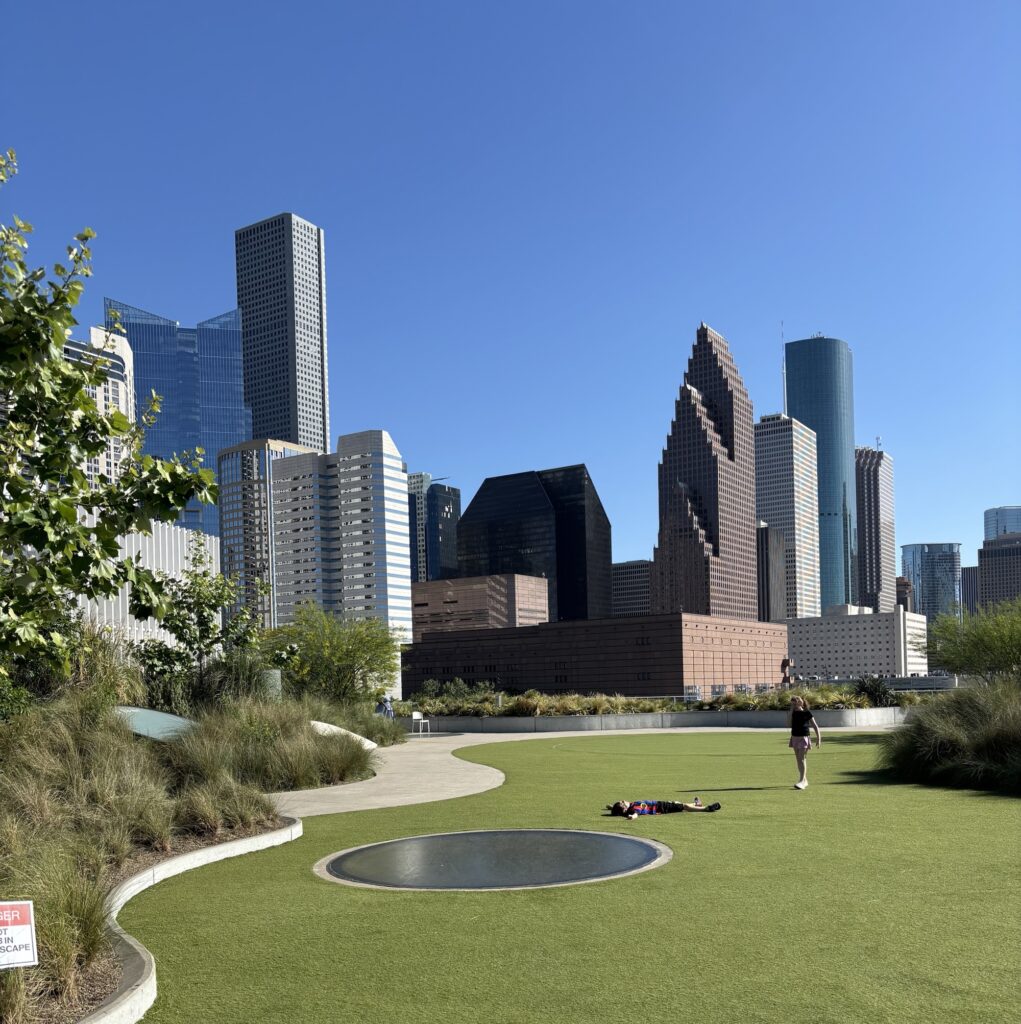 A view of downtown Houston featuring various skyscrapers under a clear blue sky, with a park area in the foreground and people enjoying the space.