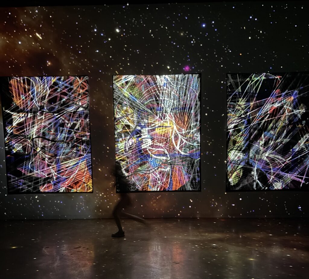 A person walking in front of large, colorful digital art displays that showcase abstract patterns and cosmic imagery in a dimly lit space. At Art Club in POST Houston.