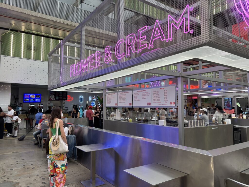 Interior of a food hall with a neon sign for 'Flower & Cream.' People are seated at tables and there are various food options visible in the background.