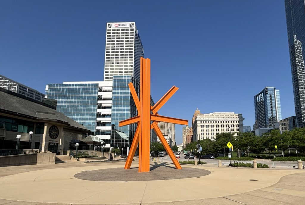 Vibrant orange modern sculpture in the downtown cityscape with skyscrapers in the background.
